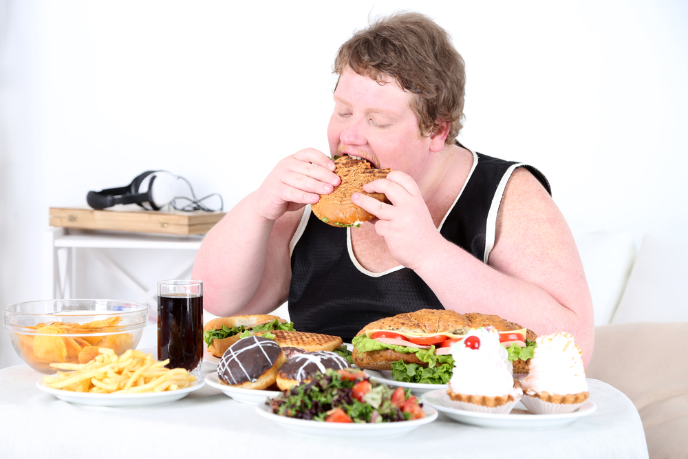 Fat man eating a lot of unhealthy food, on home interior background  