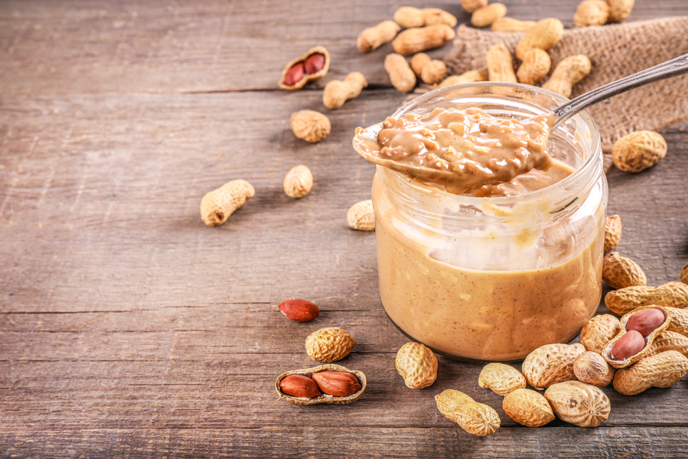 Glass jar and spoon full of crunchy peanut butter with peanuts in nutshell nearby on wooden background. Copy space.