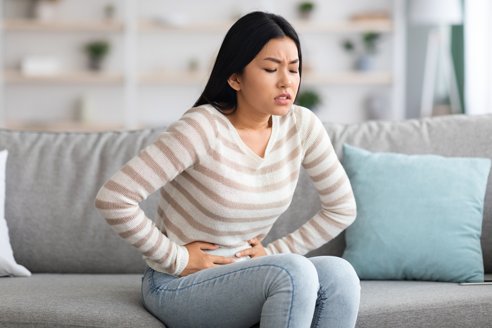 Stomach Ache. Sick Asian Woman Suffering From Acute Abdominal Pain At Home, Upset Korean Female Having Menstrual Pain Or Problems With Digestion, Sitting On Couch And Touching Belly, Free Space