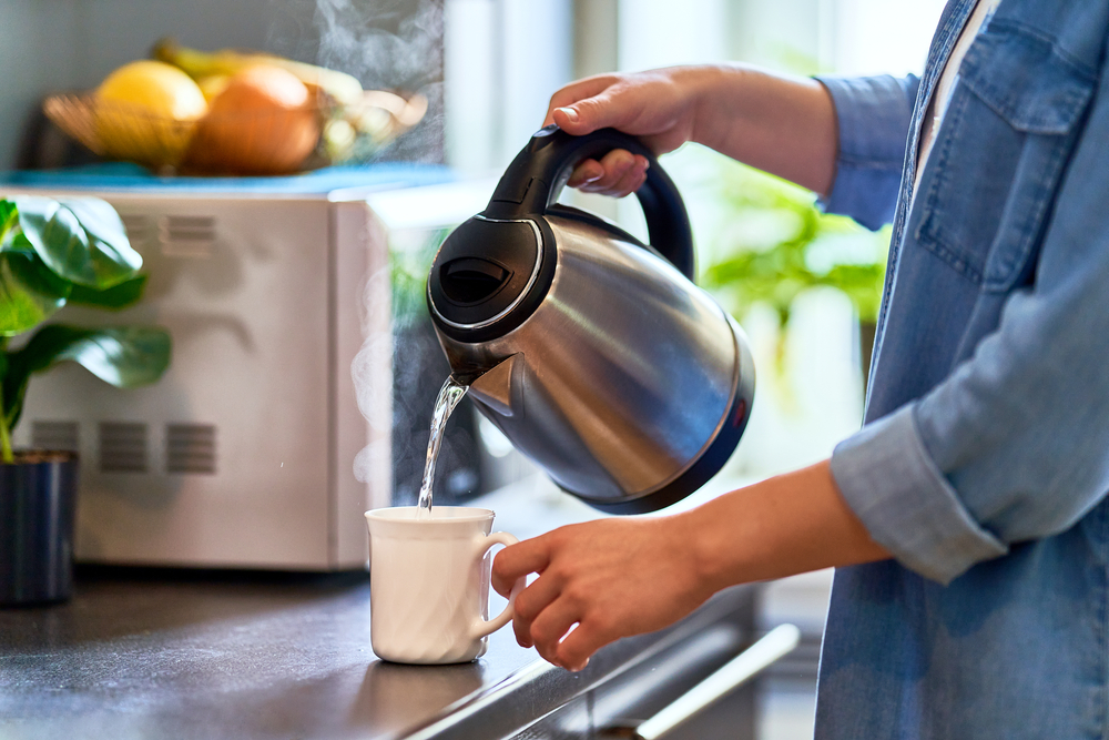 Female hands pouring boiling water from a modern metal stainless kettle in a glass cup for brewing tea in the kitchen at home