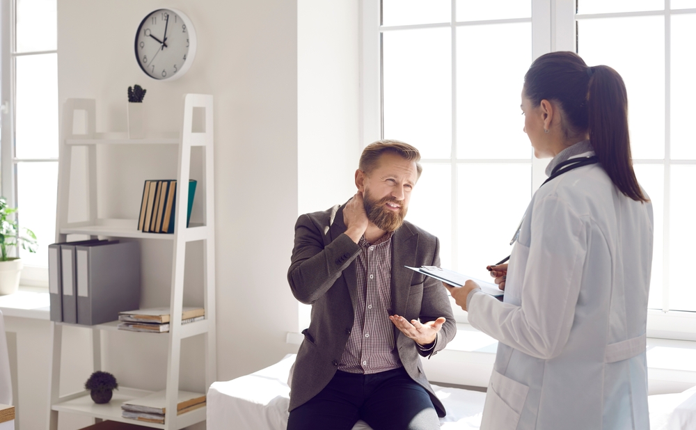 Male patient talking to doctor about pain in the nape of his neck. Female general practitioner listening to man who can't stretch stiff tight injured neck due to severe fibromyalgia or muscle strain
