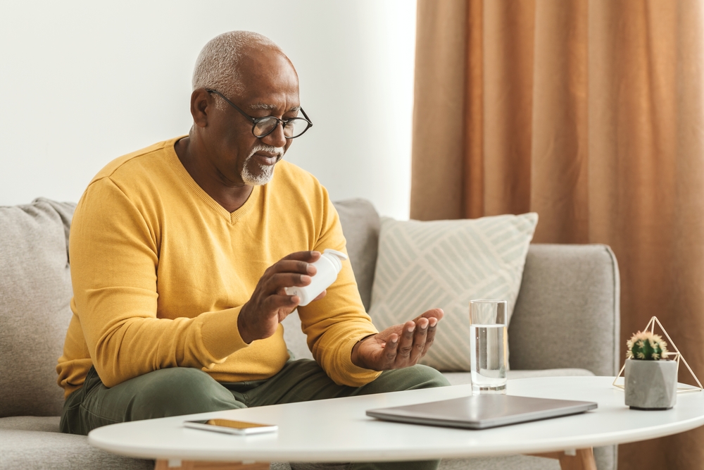 Mature Black Man Taking Medication Pill Putting Medicine On Hand Sitting Near Table With Laptop And Glass Of Water At Home. Medical Treatment, Supplements And Vitamins In Senior Age