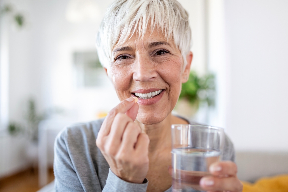 Head shot portrait happy woman holds pill glass of water, takes daily medicine vitamin D, omega 3 supplements, skin hair nail strengthen and beauty, medication for health care concept
