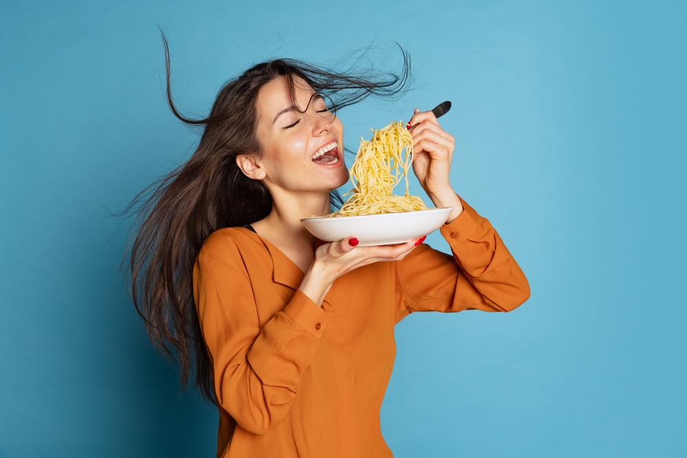Delicious taste. Beautiful young girl eating delicious Italian pasta isolated on blue studio background. Holidays, traditions, food, popularity, cafe, love. Healthy carbohydrates. Copy space for ad