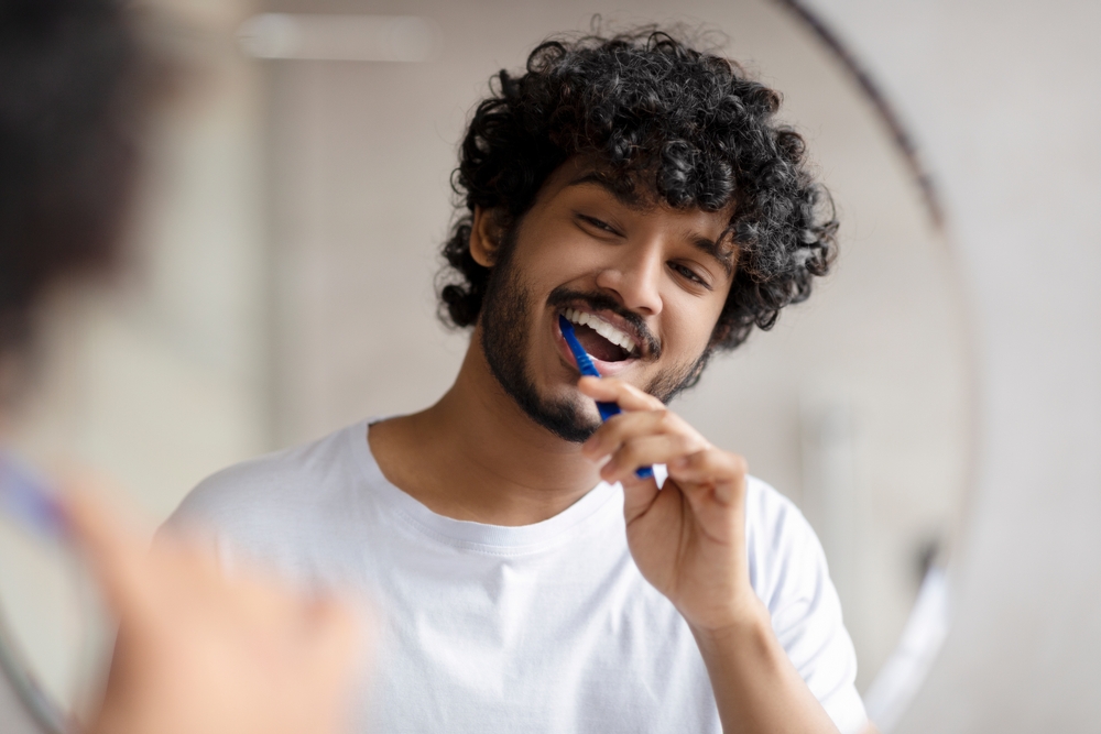 Oral care concept. Young indian man cleaning teeth with toothbrush, smiling to his reflection in mirror, doing toothcare hygiene routine in the morning in bathroom