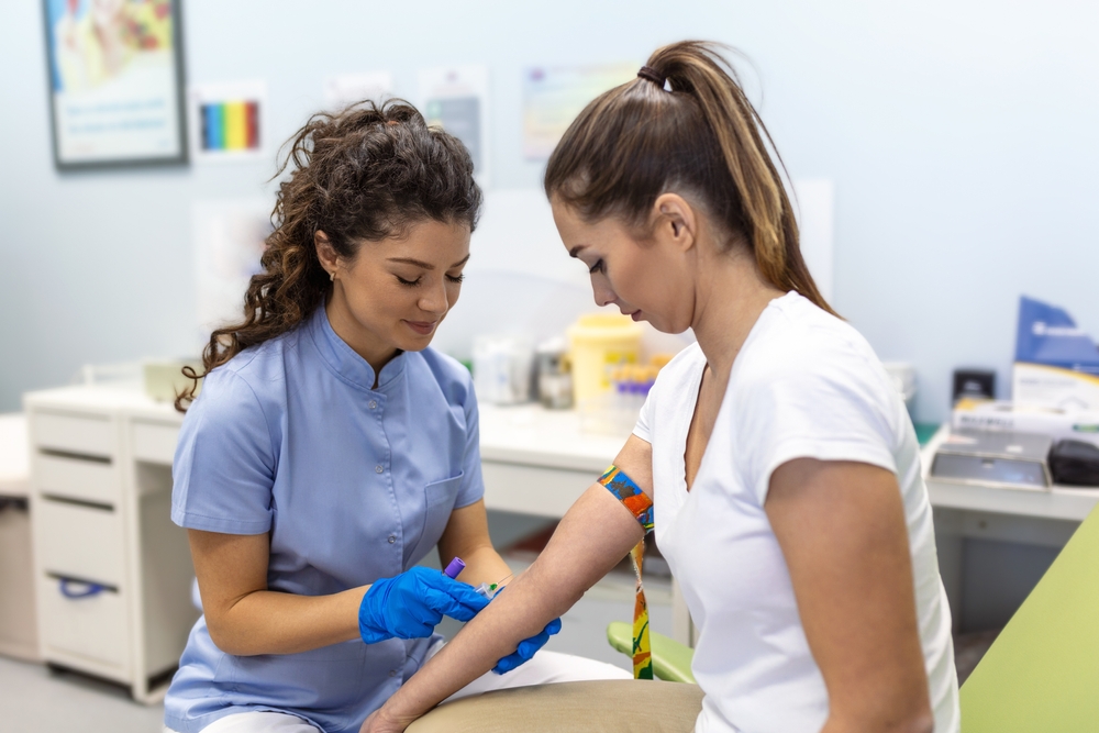 Preparation for blood test with pretty young woman by female doctor medical uniform on the table in white bright room. Nurse pierces the patient's arm vein with needle blank tube.