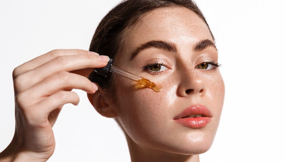 Beauty woman with freckles, naked shoulders and nude face, apply serum vitamin c, argan oil tea tree ingredients in pippette dropper under eye, treating skin for nourishing and moisturizing effect