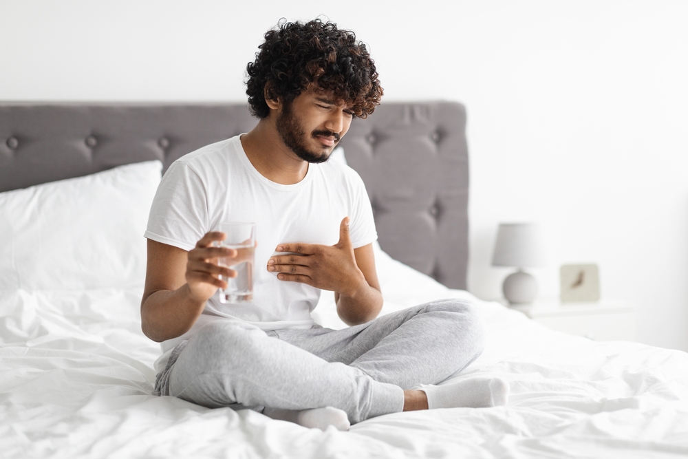 Unhappy millennial indian guy suffering from heartburn, waking up in morning, sitting on bed and holding glass of water, wearing pajamas, copy space. Acid reflux, pain in stomach concept