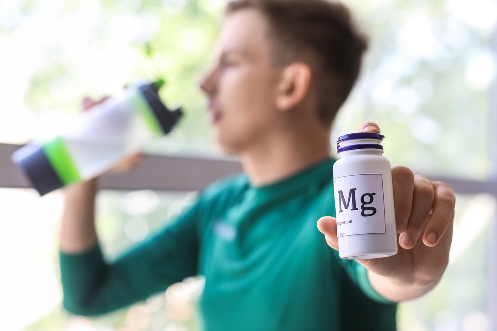 Young man with bottle of magnesium pills drinking water near window, closeup