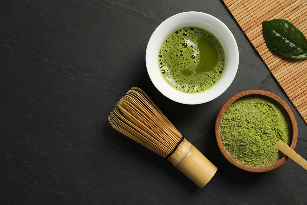 Cup of fresh matcha tea, green powder and bamboo whisk on black table, flat lay. Space for text