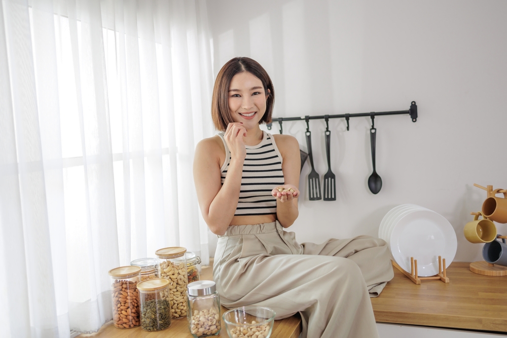 Engaging in a mindful eating moment, an Asian woman seated on a kitchen counter smiles while holding pistachios, with almonds and cashews nearby
