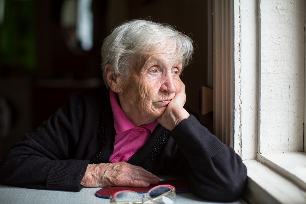 An elderly woman sadly looking out the window. Melancholy and depressed.
