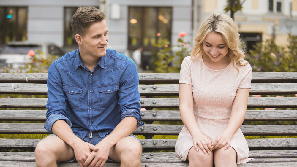 Shy blonde girl smiling, attractive guy flirting with beautiful woman on bench