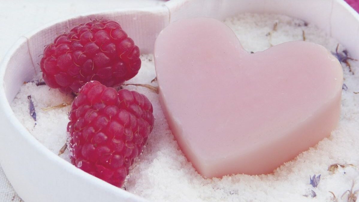 Heart shaped soap, bath salts, and raspberries in a dish. Purple flowers in the background.