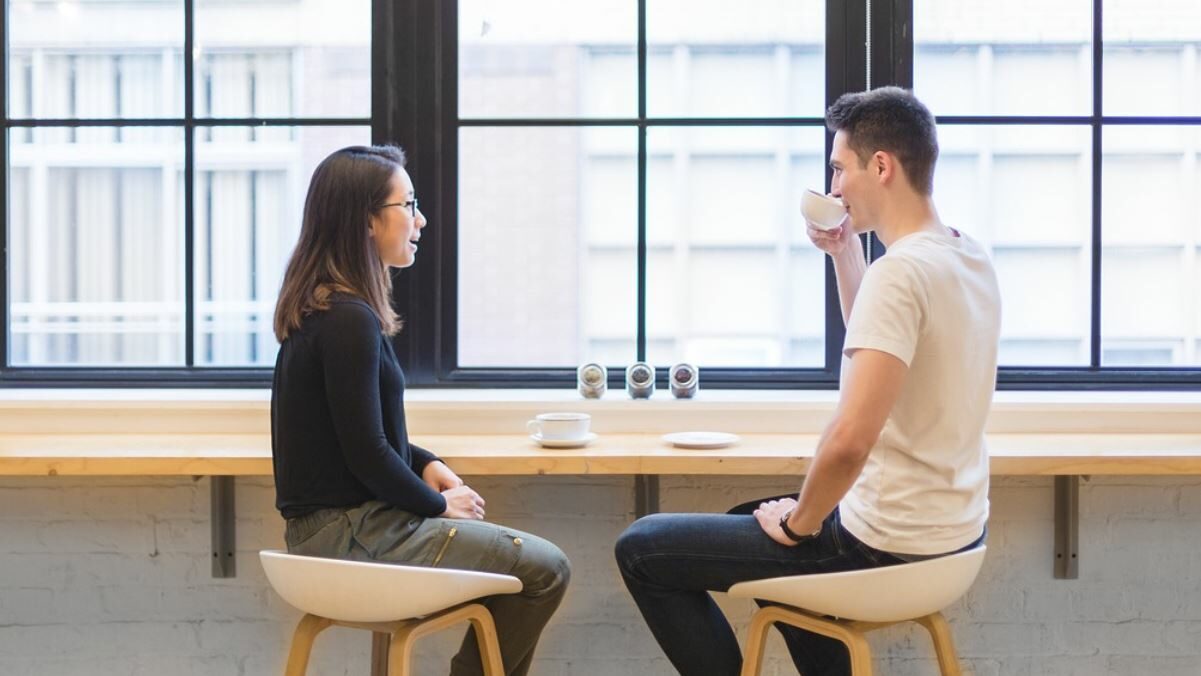 A man and woman drinking coffee and talking. Paned windows in the background.