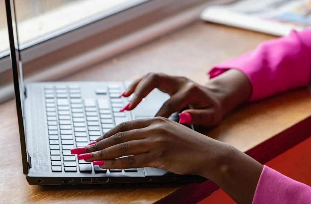 Hands with pink nails typing on a computer. Wooden desk in the background. 