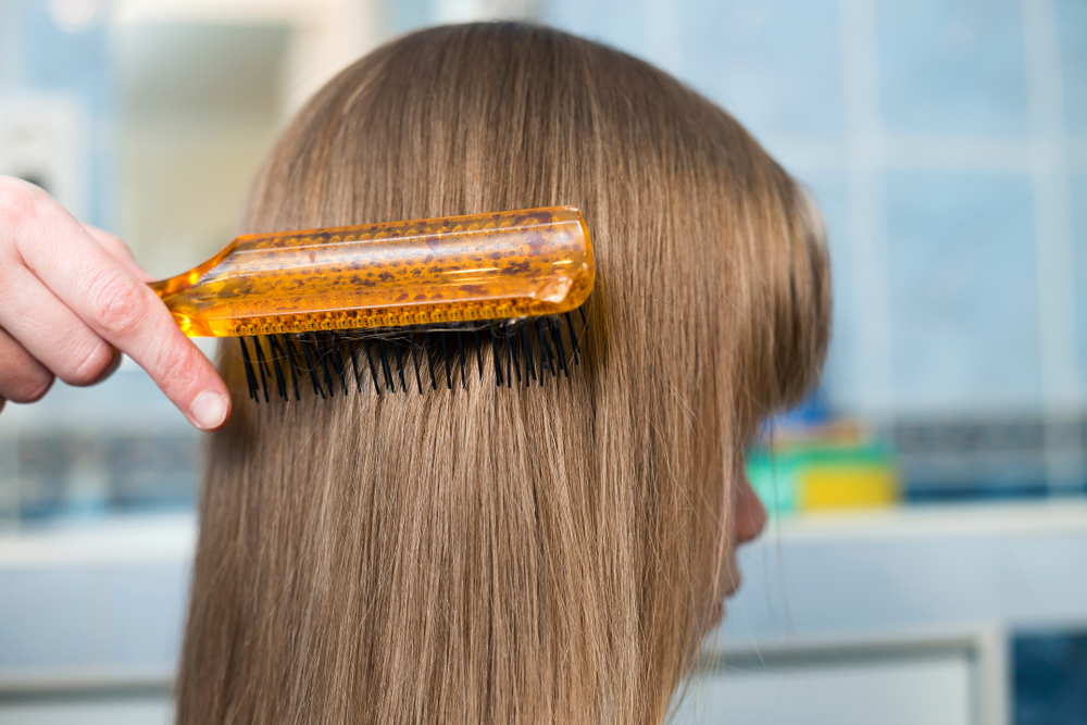 Mother hand with brush combing long fair hair of cute child girl after bath on blurred interior background.