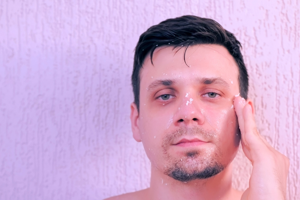 Portrait of young handsome man is applying fresh homemade coconut mask and scrub on the face. Natural organic cosmetics for skin care.