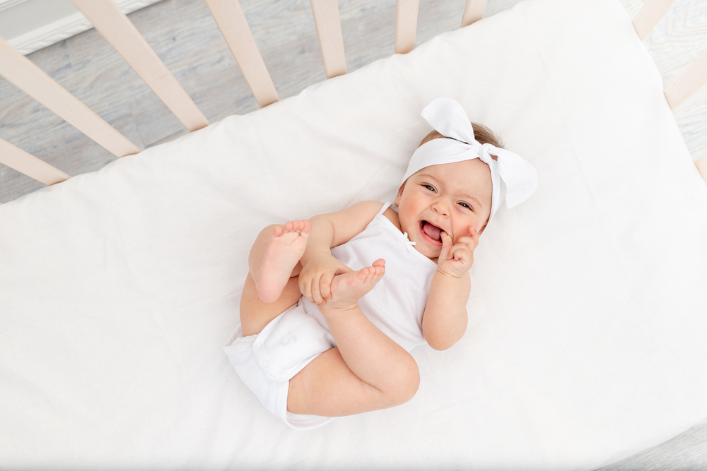 baby girl 6 months old lies in a crib in the nursery with white clothes on her back and laughs, looks at the camera, baby's morning, baby products concept