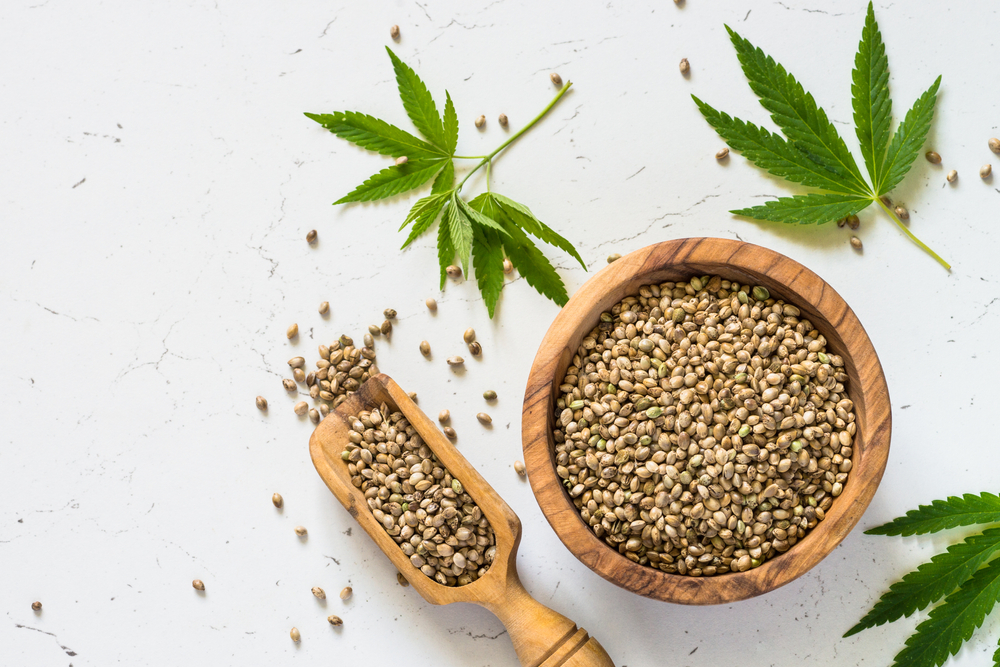 Cannabis seeds in wooden bowl and cannabis leaves at white table. Top view with copy space.