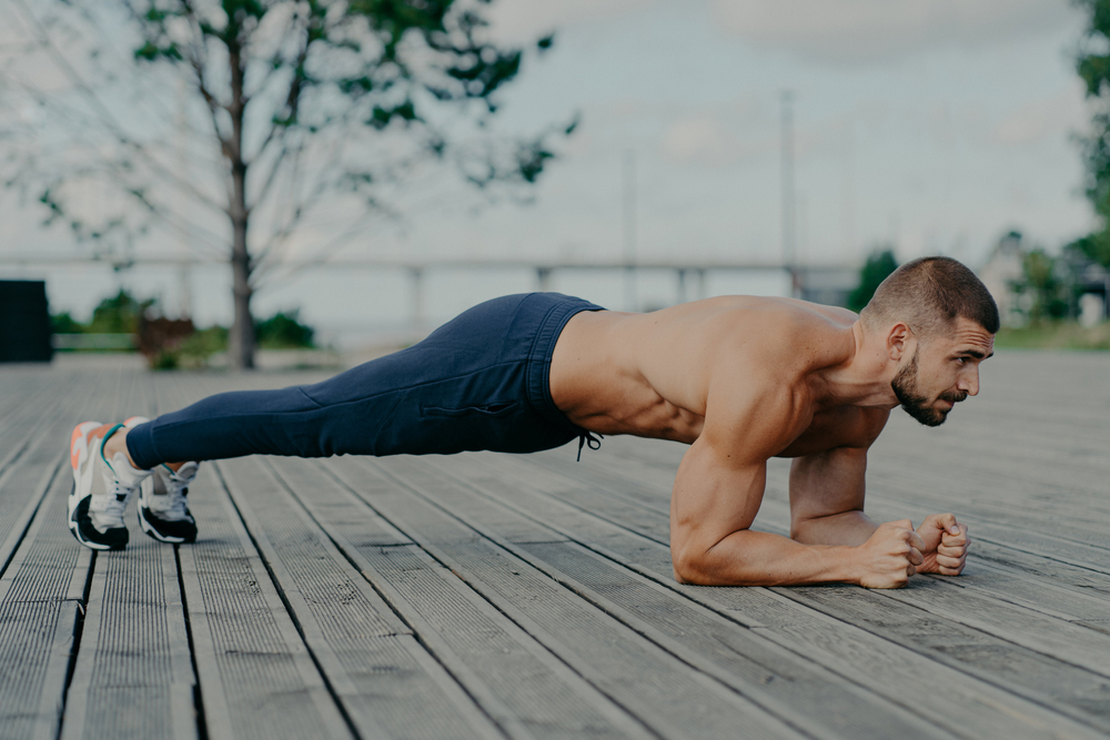 Motivated male athlete does abs exercise, enjoys bodybuilding training outdoor and stands in plank. Strong muscular European adult man has fitness workout. People and healthy lifestyle concept