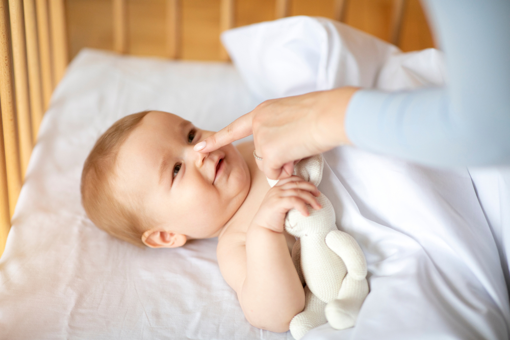 Smiling little kid laying in wooden baby bed under blanket, holding toy, mother finger touching happy kid nose, unrecognizable loving mom playing with awaken cute toddler, closeup