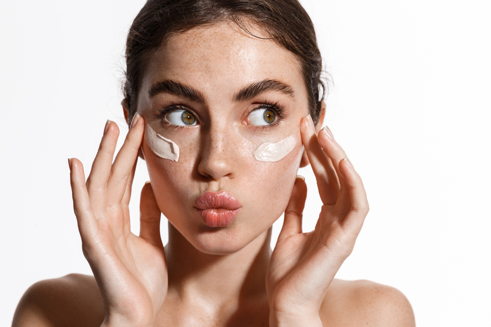 Close up beauty girl with freckles and thick eyebrows, applying moisturizing skincare cream, lotion or mask for skin lifting and anti-aging detoxifying effect, white background.