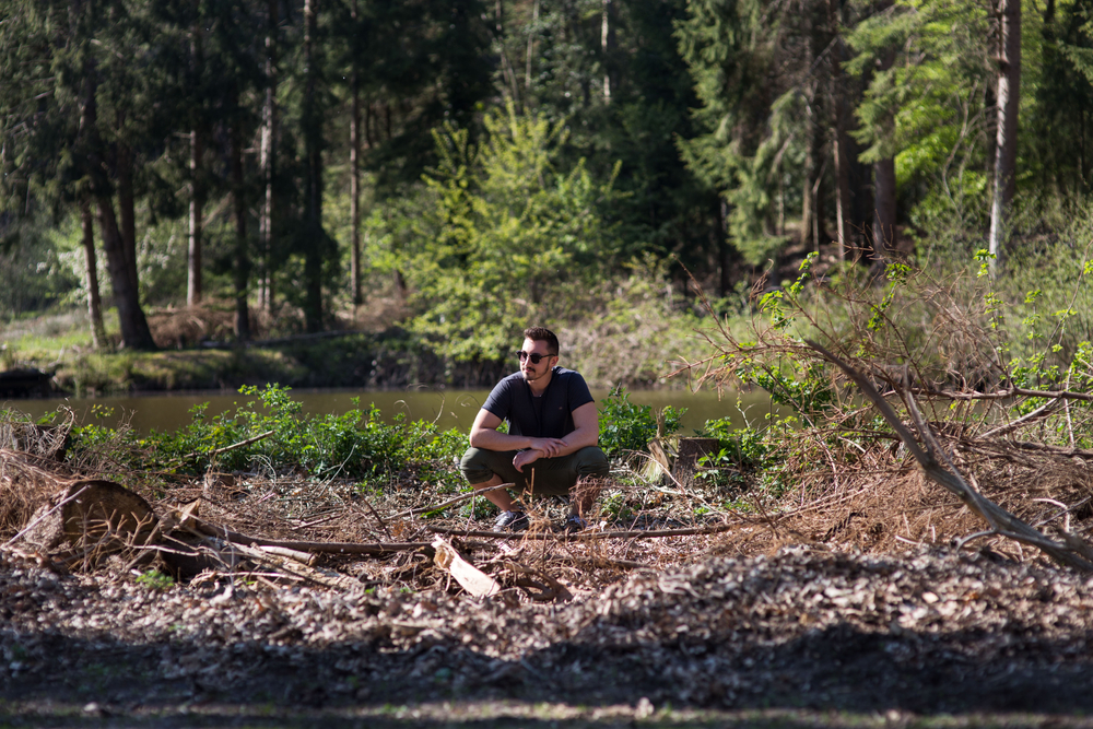 Good looking man sitting squatting very relaxed in the forest from the lake, looks to the side, He enjoys nature, thinks, loves to be free, loves nature and forest.