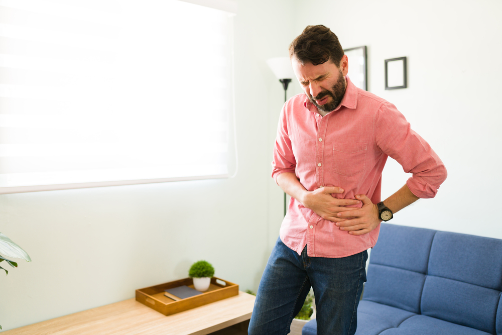 Suffering man in a lot of pain due to gallstones after standing from the couch. Young man with a gallbladder disease touching a side of his stomach