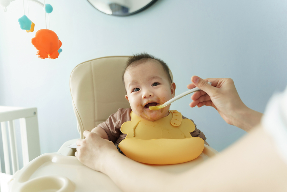 Happy Smiling Asian Little cute baby boy wearing yellow bib enjoying eating porridge or pureed food while sitting on high chair at home. Cheerful Lovely Mom feeding her son with baby spoon. BeH3althy
