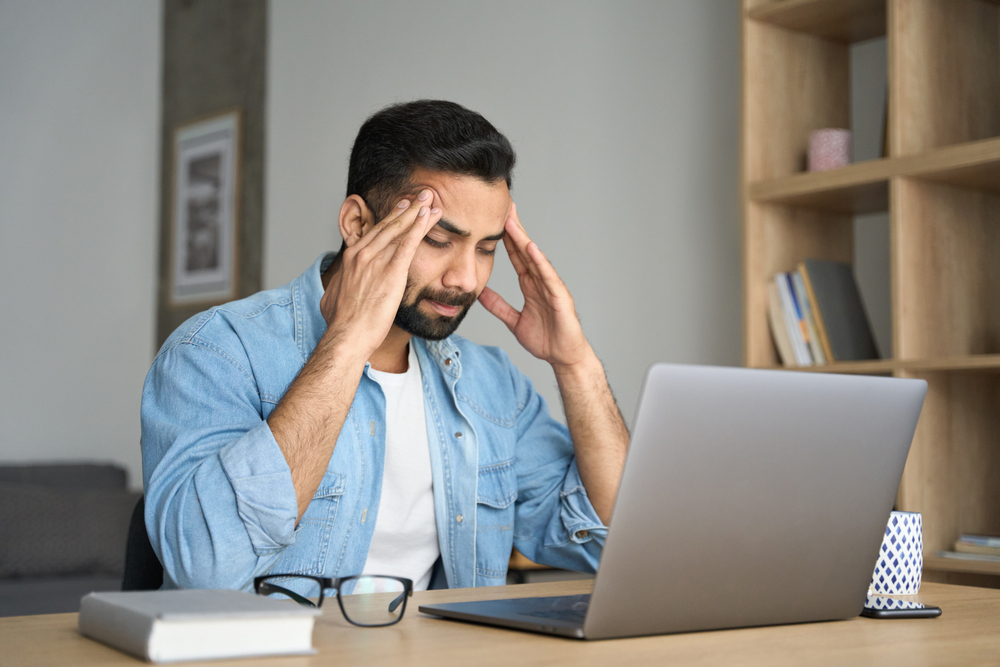 Young indian exhausted business man massaging temples suffering from headache in modern home office with laptop on desk. Overworked burnout academic Hispanic student feeling migraine head strain.