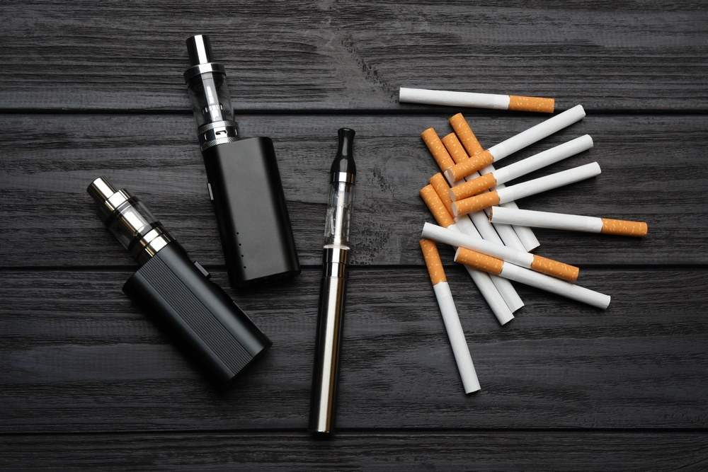 Cigarettes and different vaping devices on black wooden background, flat lay. Smoking alternative