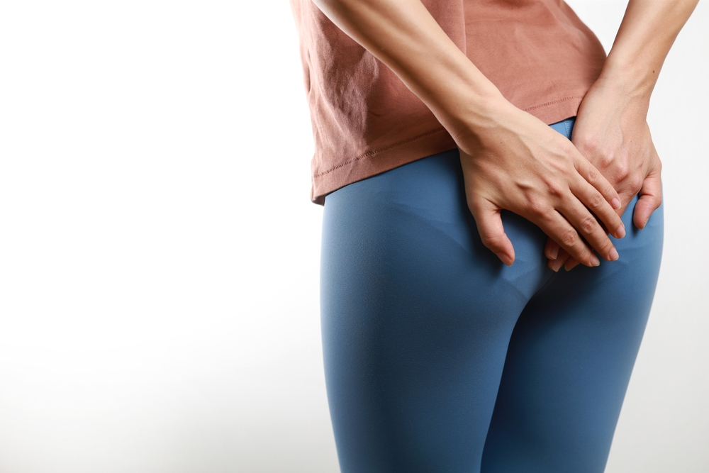 Asian woman suffering from haemorrhoids and hand holding her bottom, standing against a white background.