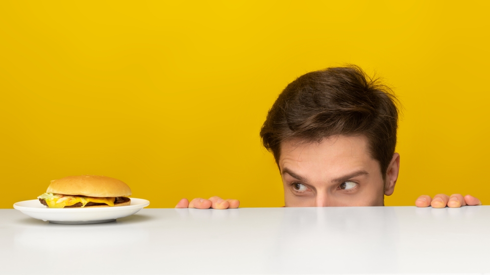 Funny Hungry Man Looking At Burger On Plate, Peeking Out Table Posing Over Yellow Orange Studio Background. Dieting, Cheat Meal And Unhealthy Junk Food, Overeating Habit Concept. Panorama Banner