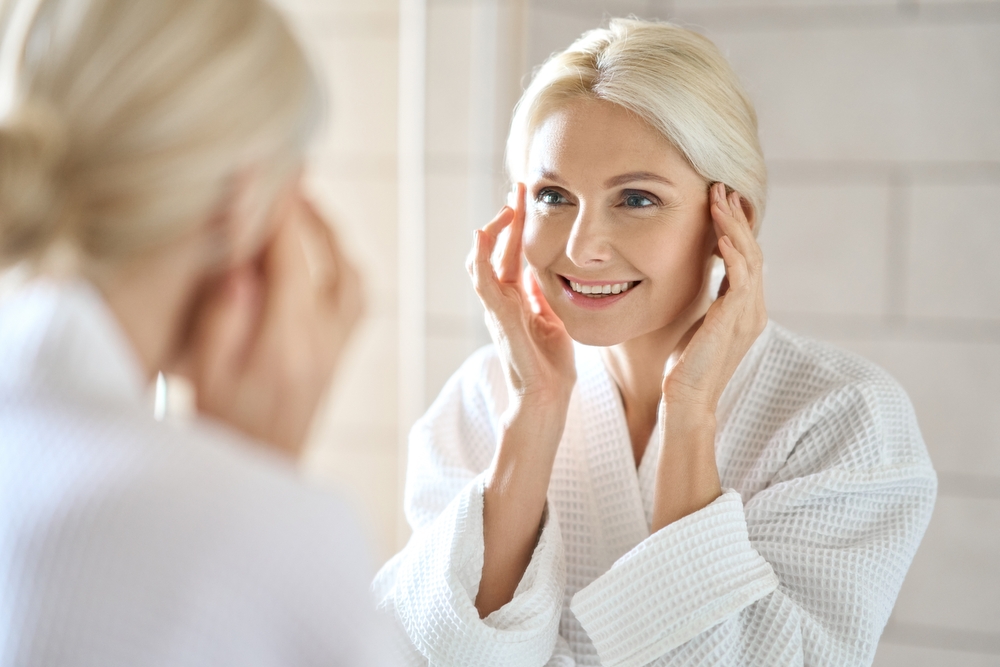 Gorgeous mid age adult 50 years old blonde woman standing in bathroom after shower touching face, looking at reflection in mirror smiling doing morning beauty routine. Older dry skin care concept.