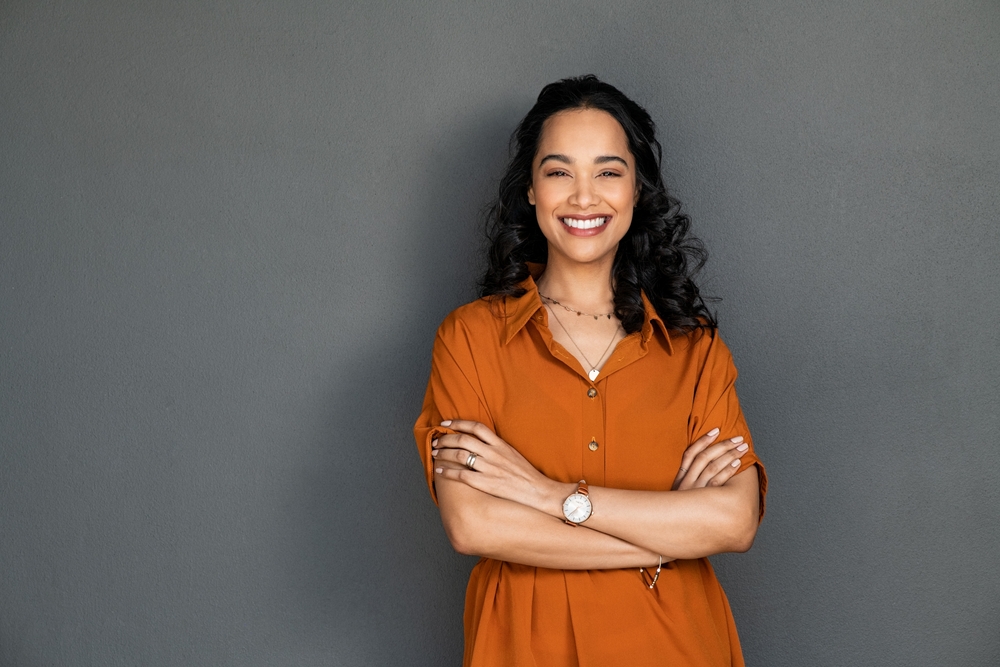 Portrait of a young latin woman with pleasant smile and crossed arms isolated on grey wall with copy space. Beautiful girl with folded arms looking at camera against grey wall. Cheerful hispanic woman