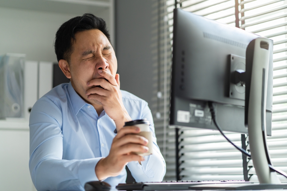 Exhausted Asain business man using laptop computer working in office. Young employee male worker sitting on table feel sleepy and yawning tired from overwork job and drinking hot coffee in workplace.