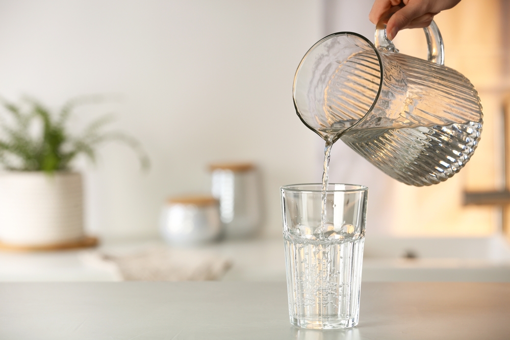 Woman pouring water from jug into glass at white table in kitchen, closeup. Space for text