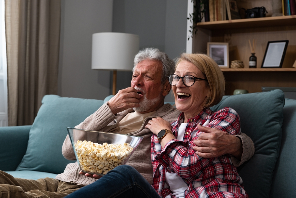 Laughing aged couple, man and woman watching tv, comedy show or movie and eating popcorn snack, sitting on cozy couch at home, mature family, man and woman enjoying free time, weekend together