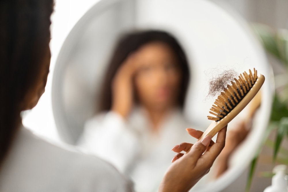 Hairloss Concept. Upset Black Woman Holding Comb Full Of Fallen Hair After Brushing, Closeup Shot Of African American Female With Brush In Hand Standing Near Mirror, Suffering Alopecia Problem