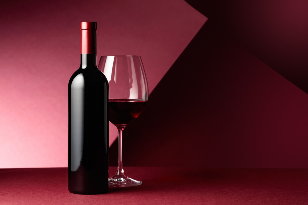 Bottle and glass of red wine on a red background. Copy space.