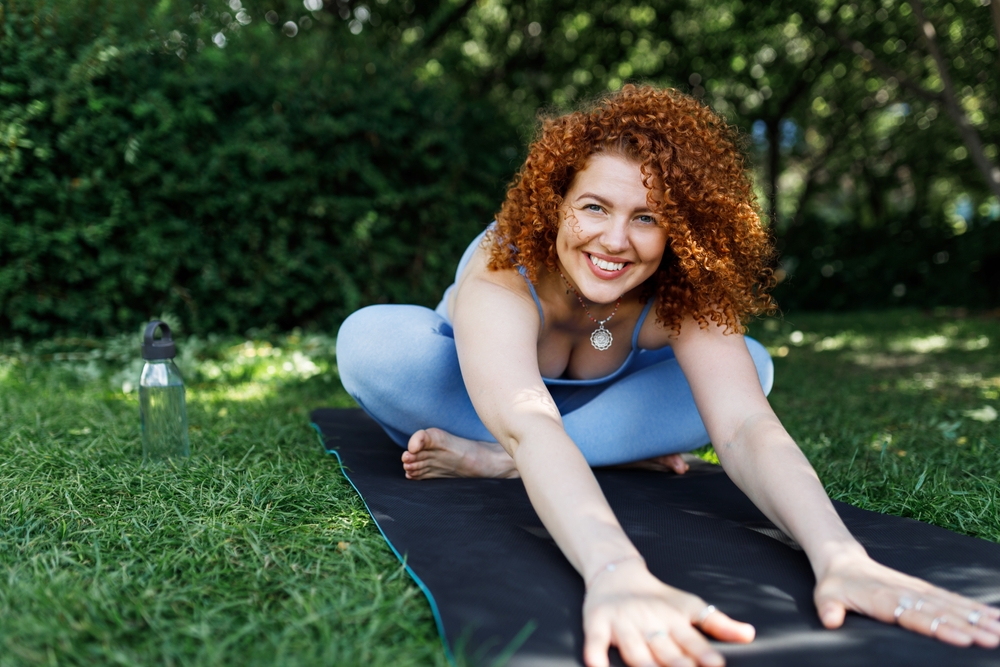 Portrait of happy pretty ginger head girl training outside in green area, sitting on mat with crossed legs, reaching hands out bending forward, smiling at camera. Yoga practice outdoor