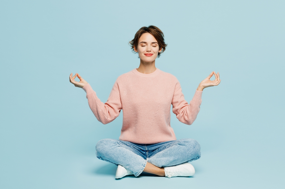 Full body young woman she wear beige knitted sweater casual clothes sit hold spreading hands in yoga om aum gesture relax meditate try to calm down isolated on plain pastel light blue cyan background