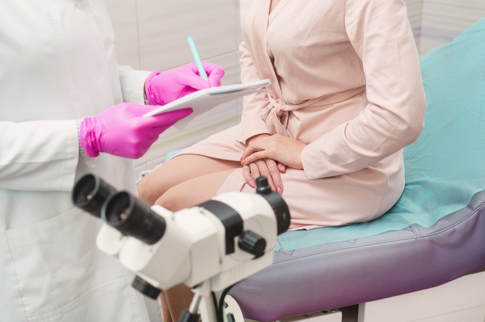 A young woman in a hospital gown at a gynecologist's appointment. A gynecologist wearing pink gloves writes a prescription to a patient. Woman on gynecological chair closeup unrecognizable colposcopy