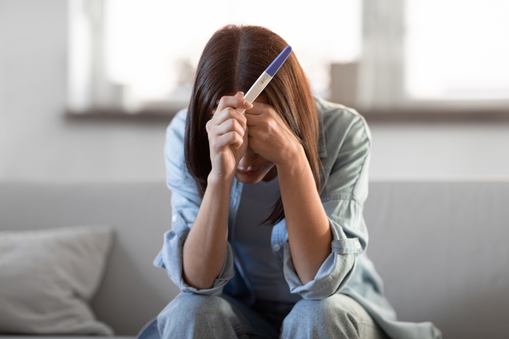Unrecognizable young woman holding pregnancy test with negative result, covering face in despair sitting at home. Maternal health problems and infertility challenges concept