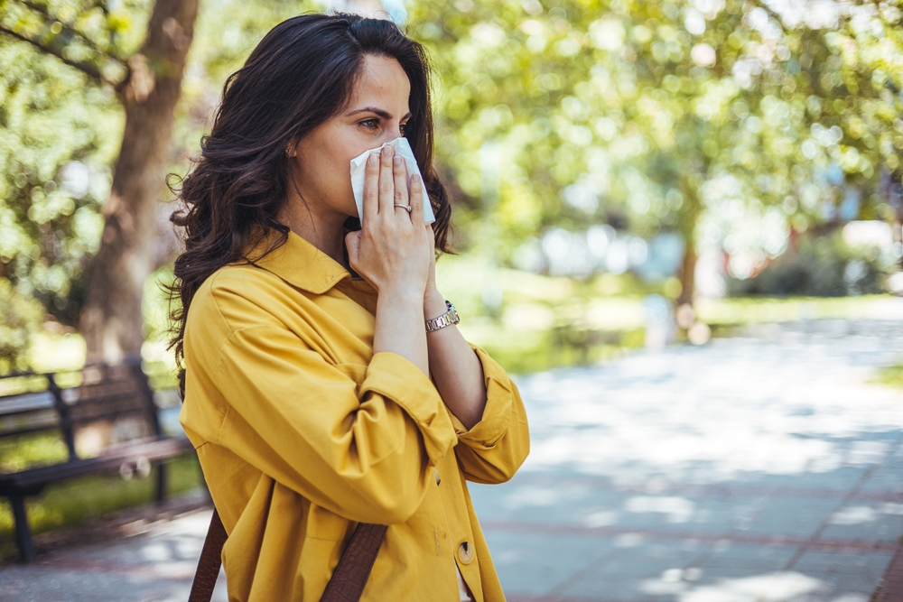 Portrait of unhealthy cute female in yellow top with napkin blowing nose, looks to the source of the allergy, place for advertising. Rhinitis, cold, allergy concept. Pollen allergy symptoms