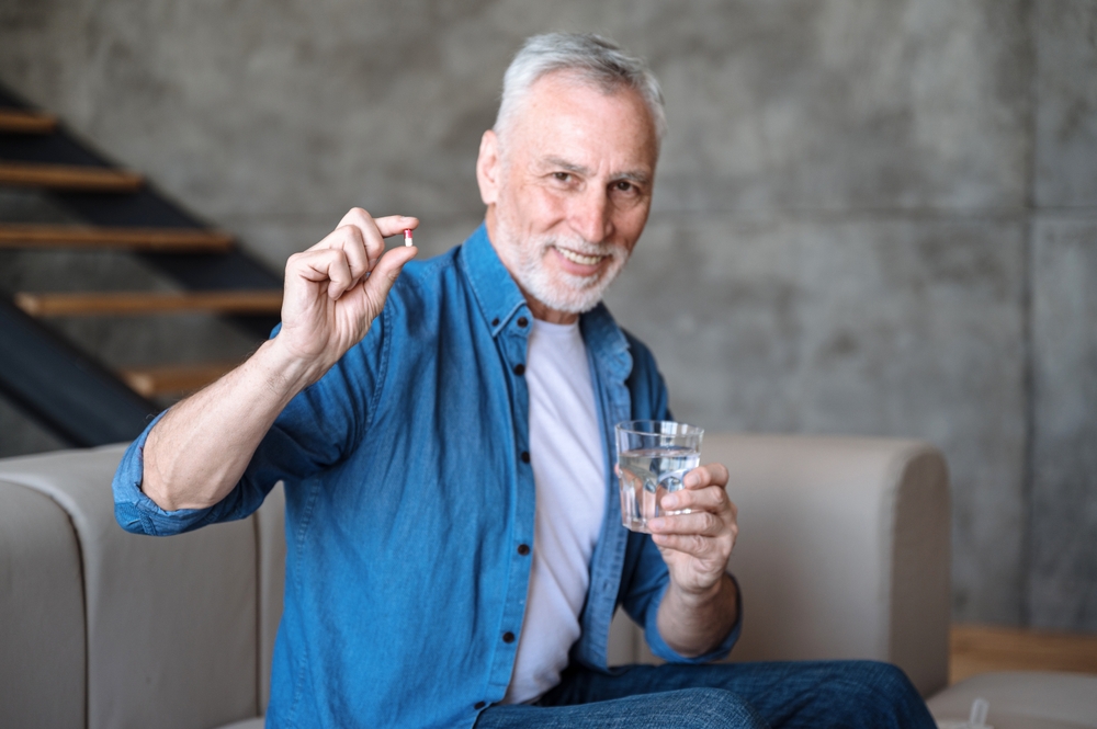 Happy smiling senior man taking supplements, daily vitamins for healthy lifestyle. Male holding tablet and glass with water. Intaking capsule with omega3, vitamin D
