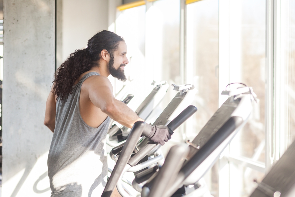 Athletic man with long curly hair wearing sportswear engaging in cardio warm-up by jogging on treadmill while gazing out the window combining fitness and scenic view. Indoor shot.