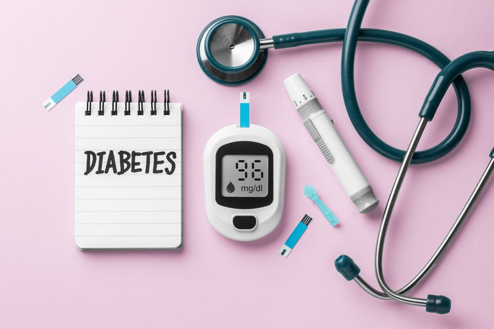 Diabetes word on notebook with blood glucose sugar meter and stethoscope on pink background, top view