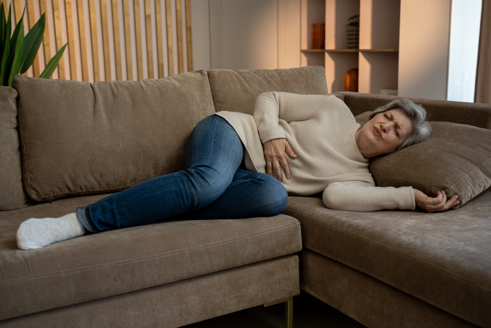Elderly woman experiencing abdominal pain while lying on a couch. She is clearly in discomfort and may be in need of medical attention.
